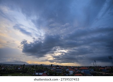 Dramatic sky with dark clouds over residential rooftops at dusk. Sunlight breaks through clouds, casting warm glow near horizon. Town below is calm, with houses and distant greenery