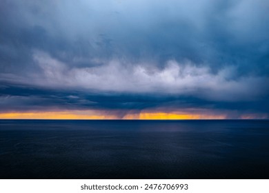 A dramatic sky with dark, stormy clouds over a tranquil sea at sunset, featuring a striking yellow and orange horizon. The image captures the contrast between the calm water and the turbulent sky.