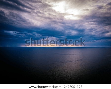 A dramatic sky with dark, stormy clouds over a tranquil sea at sunset, featuring a striking yellow and orange horizon. The image captures the contrast between the calm water and the turbulent sky.