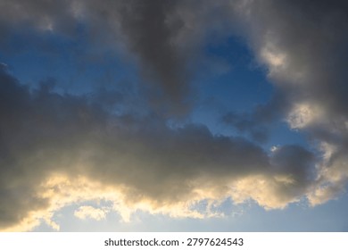 A dramatic sky featuring a combination of dark dense clouds and bright breaks in the cloud cover. The lighting creates contrast between the gray storm clouds and golden glow along the edges reminiscent of sunset or sunrise. Atmospheric skyscape.