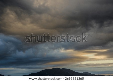 Dramatic sky filled with dark storm clouds over a mountainous landscape at sunset. The light breaking through the clouds creates a powerful contrast of warm and cool tones.