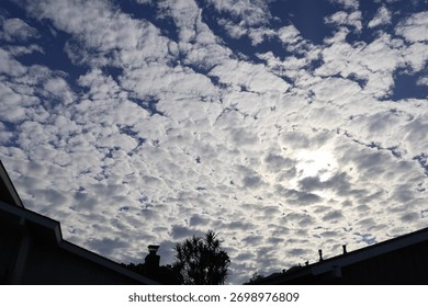 A dramatic sky filled with fluffy white clouds illuminated by the sun behind them. The sunlight filters through the dense cloud layers, creating a textured and atmospheric scene above suburban rooft