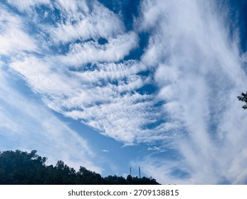 A dramatic sky filled with layered cloud streaks hangs above a forested slope, emphasizing the contrast between airy sky textures and the dense greenery of the hillside below.