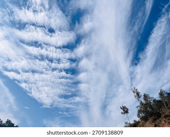 A dramatic sky filled with long sweeping cloud lines stretches over a forested hillside, capturing the striking contrast between sky textures and lush mountain greenery.