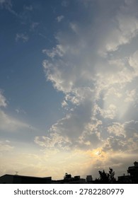 Dramatic sky with fluffy clouds and sun rays breaking through during sunset over a dark silhouette of city buildings.