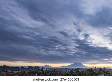 Dramatic sky looms above distant mountains and rooftops. Soft golden light breaks through darkening clouds. Village rooftops peek through dense greenery. Misty peaks stand silent under gathering storm