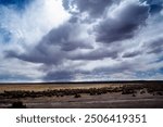 A dramatic sky looms over the arid plains of Bolivia, with dark, heavy clouds casting shadows across the golden-brown landscape. The barren terrain contrasts sharply with the turbulent sky above.