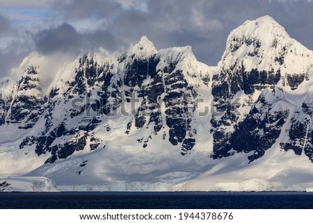 Dramatic sky over craggy peaks of a majestic frozen ridge of a mountain range of icy coastal mountains and a rock face on an ice cold cape on the mountainous and scenic Antarctic coast of Antarctica