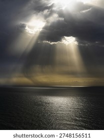 Dramatic sky with radiant sun rays piercing stormy clouds above calm sea surface, creating serene and powerful natural scene