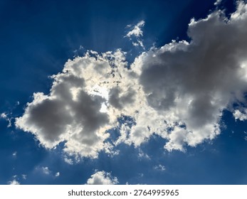 A dramatic sky scene featuring dense, billowing clouds partially covering the sun against a deep blue background. Bright sunlight filters through gaps in the clouds, creating striking crepuscular rays that spread outward in soft, radiant beams. The cloud edges glow with a silver lining effect, highlighting their texture and depth, while darker patches add contrast and dimension. This natural composition captures the interplay of light and shadow, evoking a sense of calm, power, and atmospheric beauty. Ideal for themes of nature, tranquility, weather, and inspiration, the image offers a high-resolution sky background suitable for wallpapers, digital design, and stock use. The balance of brightness and shadow enhances visual appeal, making it perfect for creative and commercial applications.