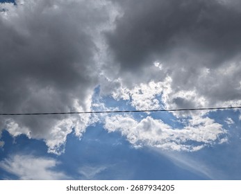 A dramatic sky scene featuring a mix of dark storm clouds and bright white clouds against a blue sky, with a power line stretching across the horizon.