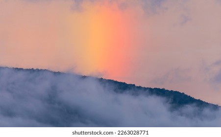 Dramatic sky shining rainbow behind the foggy mountain after raining at twilight.