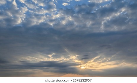 A dramatic sky with sunbeams piercing through a layer of dark clouds. The upper sky is a beautiful blue with scattered, illuminated clouds, creating a powerful scene of light and shadow.