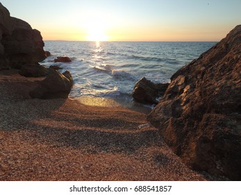 Dramatic sky: sunset beach at dusk captures shoreline during golden hour as sun reflects off water, creating shimmering trails across gentle waves. Ocean transitions from deep navy to lighter azure near rocky shore where textured stones contrast with sandy foreground. Warm hues cast across skyline create glowing ambiance at day's end while waves lap softly against coastline. This composition emphasizes vastness with wide-angle perspective focusing on detailed textures of pebbles and shells. Peaceful mood invites relaxation and reflection for wellness themes and travel imagery. Natural setting works for lifestyle content and mental health campaigns with vibrant colors.