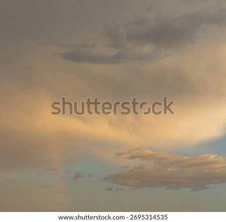 A dramatic sky at sunset or sunrise with heavy, thick clouds displaying moody gray and soft golden-peach hues. A faint, partial rainbow or sun dog (parhelion) is visible in the upper left.