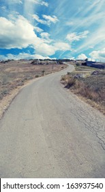 Dramatic sky: winding roadway meanders through serene terrain under vast blue skyscape dotted with scattered fluffy white clouds. Paved thoroughfare curves gently toward horizon, inviting exploration with visible cracks and slight sheen on surface. Surrounding landscape features parched earth with patches of dry turf and sparse vegetation, creating tranquil outside setting. Rustic structures with muted tones blend harmoniously into surroundings, adding quaint rural charm to this atmospheric depiction. Soft diffuse sunlight enhances warm hues of terrain below while expansive overhead provides sense of openness and freedom. This photographic scene conveys peaceful exploration narrative ideal for travel marketing, outdoor lifestyle campaigns, and environmental awareness projects. Composition frames path as leading line directing gaze toward distant skyline with weathered textures and ambient atmosphere.
