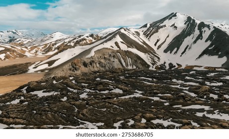 Dramatic snow-capped volcanic mountains overlooking a vast field of dark, textured lava rock under a cloudy sky