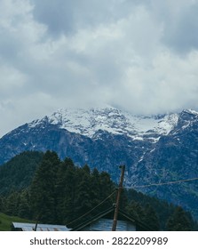 : A dramatic snow-covered mountain peak partly wrapped in cloud, viewed above a dense pine forest and a rustic tin-roofed cabin.