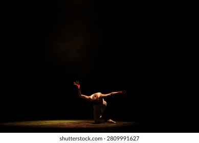 A dramatic solo dance moment featuring expressive body movement illuminated by a single theatrical spotlight against black space.