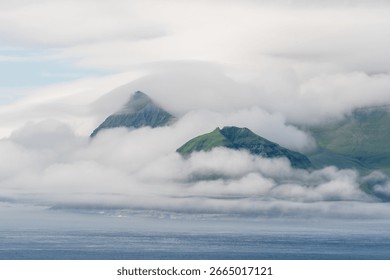 A dramatic, steep-sided green mountain rises from the ocean, shrouded in mist and clouds, capturing the wild beauty of the Faroe Islands.