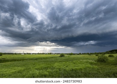 A dramatic storm cloud hangs low over a field of green grass, threatening rain. The sun peeks through the clouds, casting beams of light onto the meadow below.