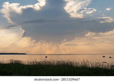 Dramatic storm cloud with visible rain shafts over a calm sea at sunset. Sun rays break through the clouds, creating a peaceful yet powerful coastal landscape.
