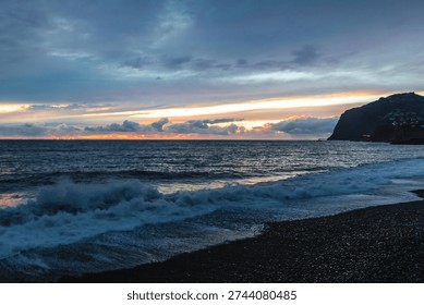 Dramatic storm clouds gather over the rugged volcanic shoreline of Madeira as Atlantic waves crash onto a dark pebble beach at dusk.



