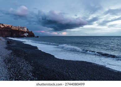 Dramatic storm clouds gather over the rugged volcanic shoreline of Madeira as Atlantic waves crash onto a dark pebble beach at dusk.



