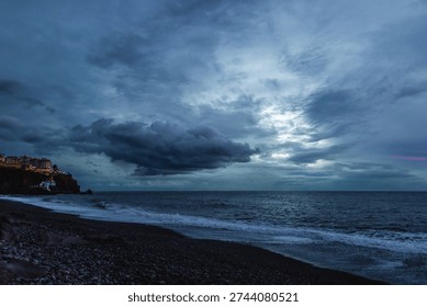 Dramatic storm clouds gather over the rugged volcanic shoreline of Madeira as Atlantic waves crash onto a dark pebble beach at dusk.



