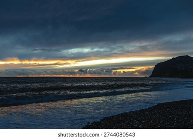Dramatic storm clouds gather over the rugged volcanic shoreline of Madeira as Atlantic waves crash onto a dark pebble beach at dusk.



