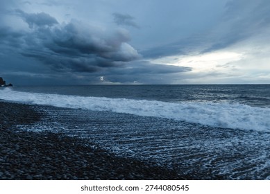 Dramatic storm clouds gather over the rugged volcanic shoreline of Madeira as Atlantic waves crash onto a dark pebble beach at dusk.



