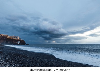 Dramatic storm clouds gather over the rugged volcanic shoreline of Madeira as Atlantic waves crash onto a dark pebble beach at dusk.



