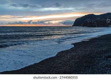 Dramatic storm clouds gather over the rugged volcanic shoreline of Madeira as Atlantic waves crash onto a dark pebble beach at dusk.



