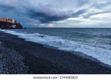 Dramatic storm clouds gather over the rugged volcanic shoreline of Madeira as Atlantic waves crash onto a dark pebble beach at dusk.




