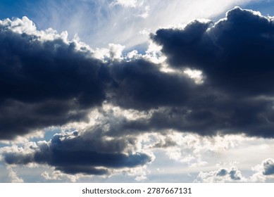 Dramatic storm clouds gathering in the bright blue sky with glowing sunlight breaking through the dark heavy masses creating a moody and atmospheric ethereal celestial weather vista