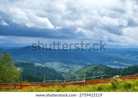 Dramatic storm clouds hang over a sweeping view of a forested mountain valley with a small village and a safety barrier in the foreground