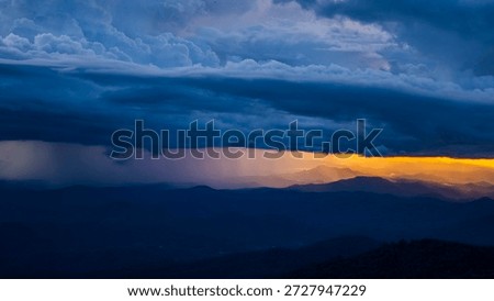 Dramatic storm clouds with heavy rain over mountain silhouette during sunset. Dark sky contrast with golden sunlight background.