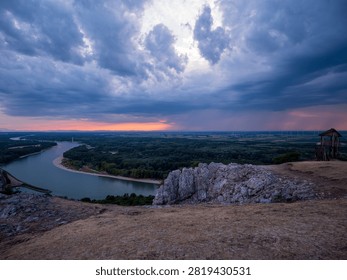 Dramatic storm clouds over the Danube River viewed from Braunsberg Hill near Hainburg an der Donau, Austria, with rocky foreground and lush floodplain forest at sunset.