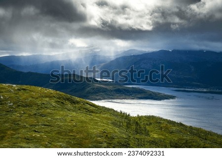 Dramatic storm clouds over the mountains and the Bergsfjorden fjord on Senja Island, with rain showers and ethereal rays of light piercing the tempest, as seen from the trail to Husfjellet Mountain.