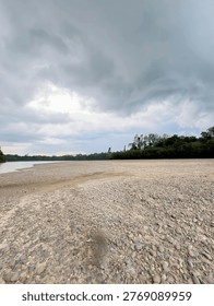 Dramatic Storm Clouds Over Rocky River Bank 

A wide landscape of a pebble-covered riverbank under a heavy, dramatic grey sky. Lush green tropical trees line the horizon of the peaceful water.
