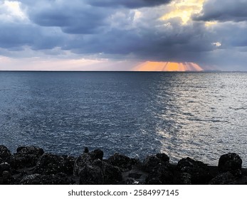 Dramatic storm clouds over a vast ocean, with golden sun rays piercing through the dark sky. The moody seascape contrasts deep blue waters with warm light breaking through in the distance.