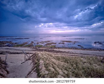 Dramatic storm clouds rolling over the coastline at Pointe de Grignon, Ars-en-Re, Ile de Re, France, with a sandy dune path leading to calm tidal pools at dusk