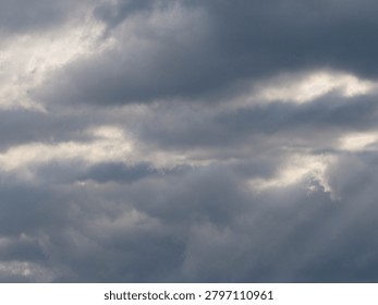Dramatic Storm Clouds with Sun Rays Breaking Through Overcast Sky. A dramatic sky filled with dark gray storm clouds and textured overcast formations. Sunbeams break through gaps in the cloud cover, creating atmospheric rays of light and a moody weather scene. Natural cloudscape background with changing weather conditions, suitable for themes of nature, climate, hope, spirituality, forecast, and environmental concepts.