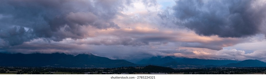 Dramatic storm clouds sweep across the Chilliwack mountain range at sunset, forming a moody British Columbia panorama.