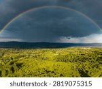 Dramatic storm sky with a vibrant full rainbow arch over green fields and rural landscape. Aerial drone photography.