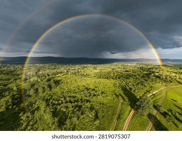 Dramatic storm sky with a vibrant full rainbow arch over green fields and rural landscape. Aerial drone photography.