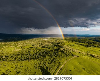 Dramatic storm sky with a vibrant full rainbow arch over green fields and rural landscape. Aerial drone photography.