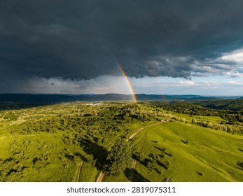 Dramatic storm sky with a vibrant full rainbow arch over green fields and rural landscape. Aerial drone photography.