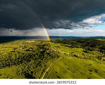 Dramatic storm sky with a vibrant full rainbow arch over green fields and rural landscape. Aerial drone photography.