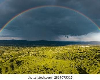 Dramatic storm sky with a vibrant full rainbow arch over green fields and rural landscape. Aerial drone photography.